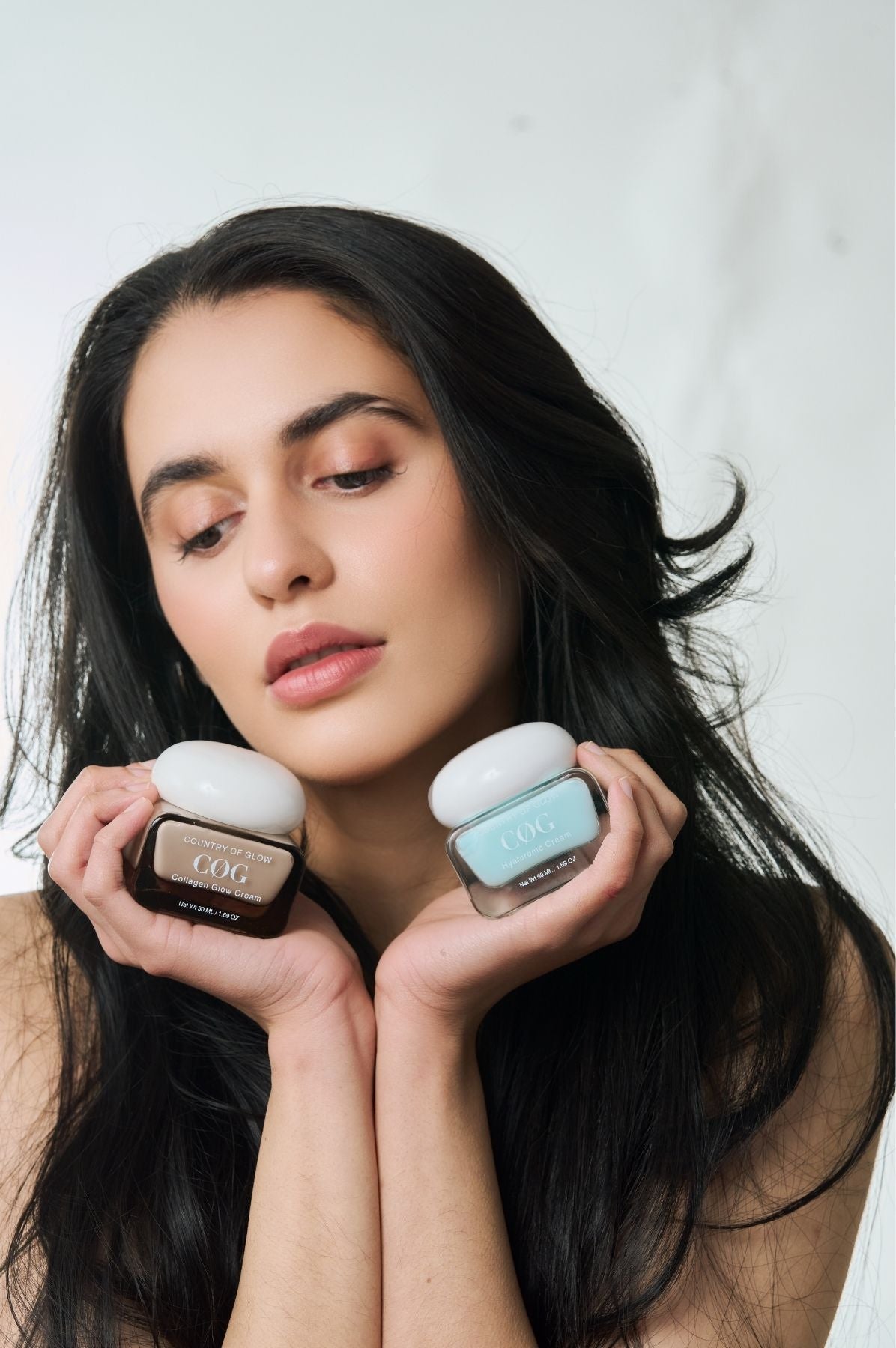 A woman with long dark hair holds two cosmetic jars near her chin against a light neutral background. One jar is dark brown with a white lid and the other is light blue-gray with a white lid.