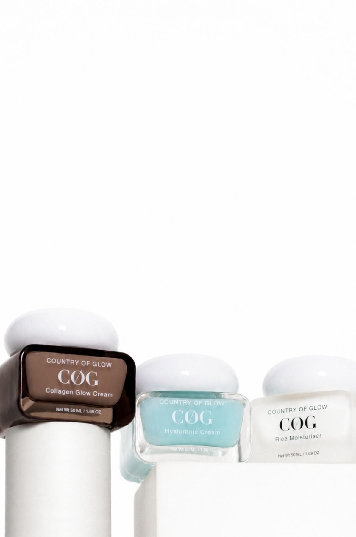 Two COG skincare jars on a textured stone surface with dramatic side lighting. One jar is frosted light blue with a gray lid, and the other is white with a gray lid.
