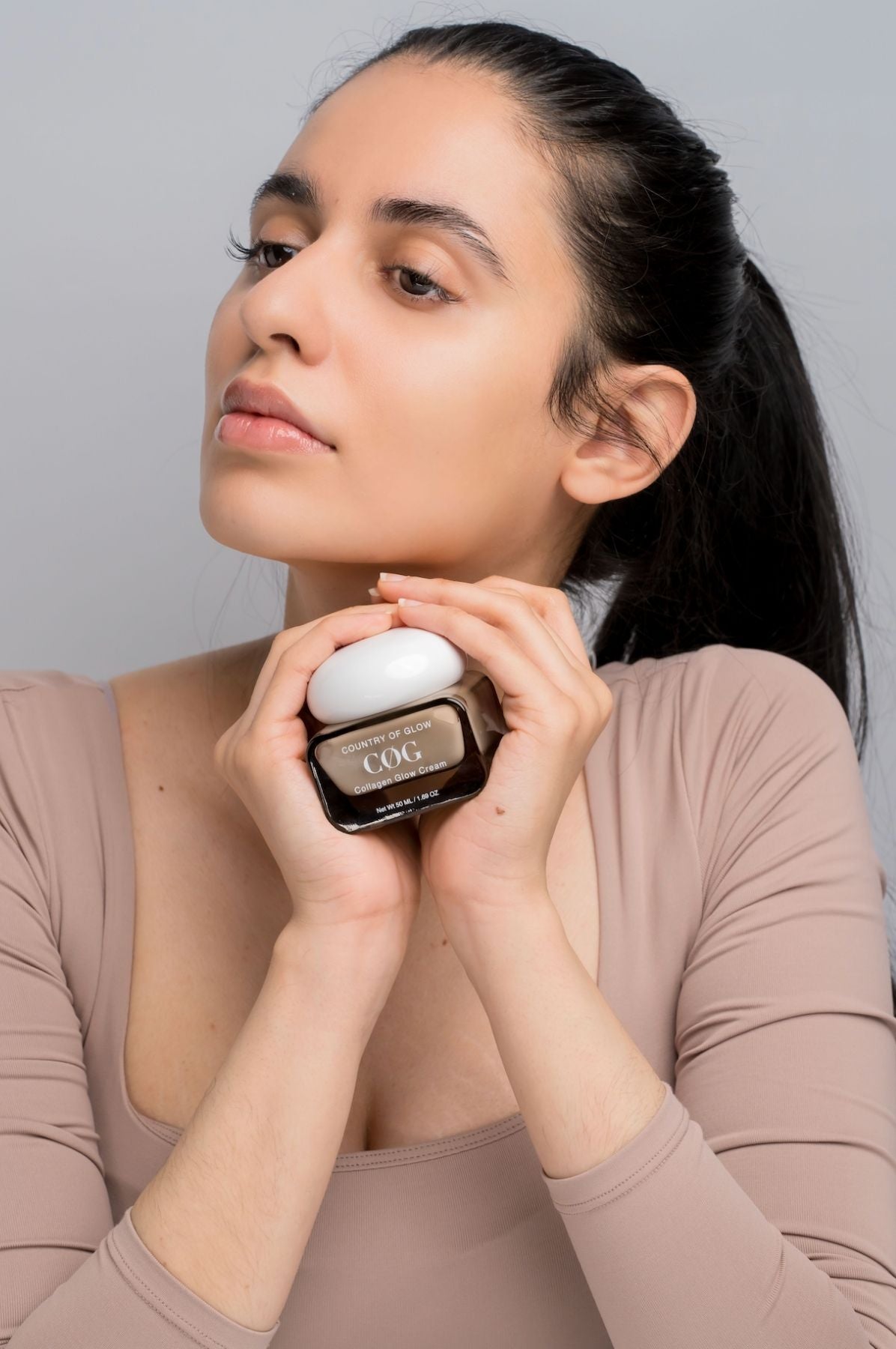 A woman with dark hair in a ponytail holds a cosmetic jar near her neck. The jar has a dark brown glass body with a white lid and a label reading 'COG'.