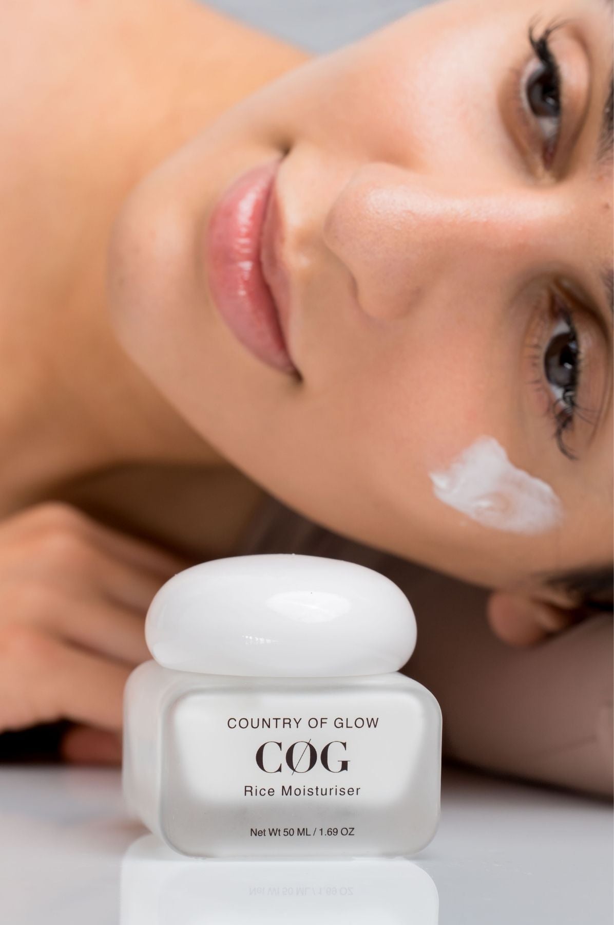 A close-up of a woman lying down with a white cream applied to her cheek, with a jar of Country of Glow COG Rice Moisturiser in the foreground on a reflective white surface.