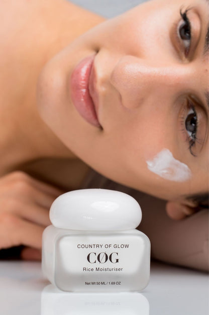 A close-up of a woman lying down with a white cream applied to her cheek, with a jar of Country of Glow COG Rice Moisturiser in the foreground on a reflective white surface.